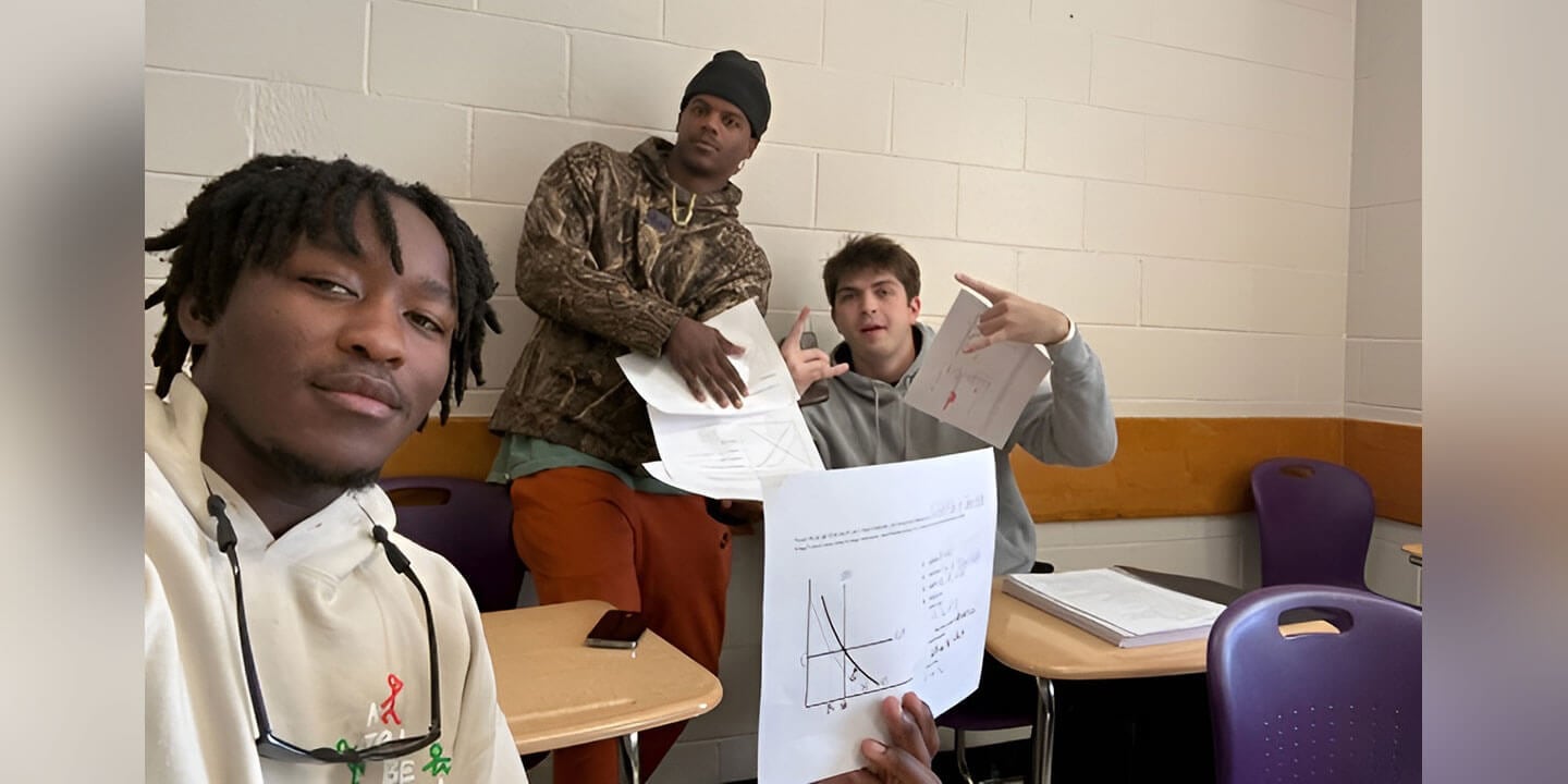 Three students seated in a classroom, all three are holding papers; one shows a math diagram on it.