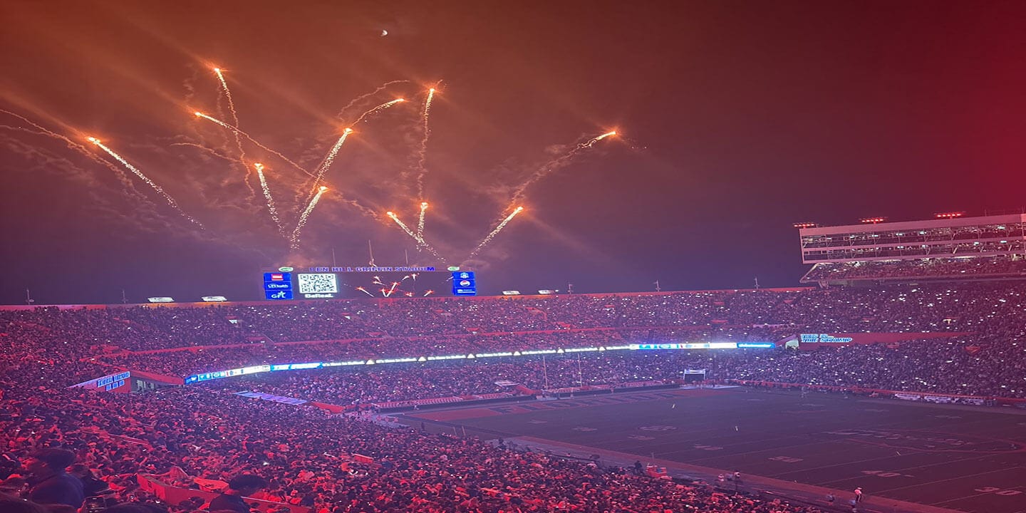 A college football stadium filled to capacity at night with fireworks exploding over the end zone.