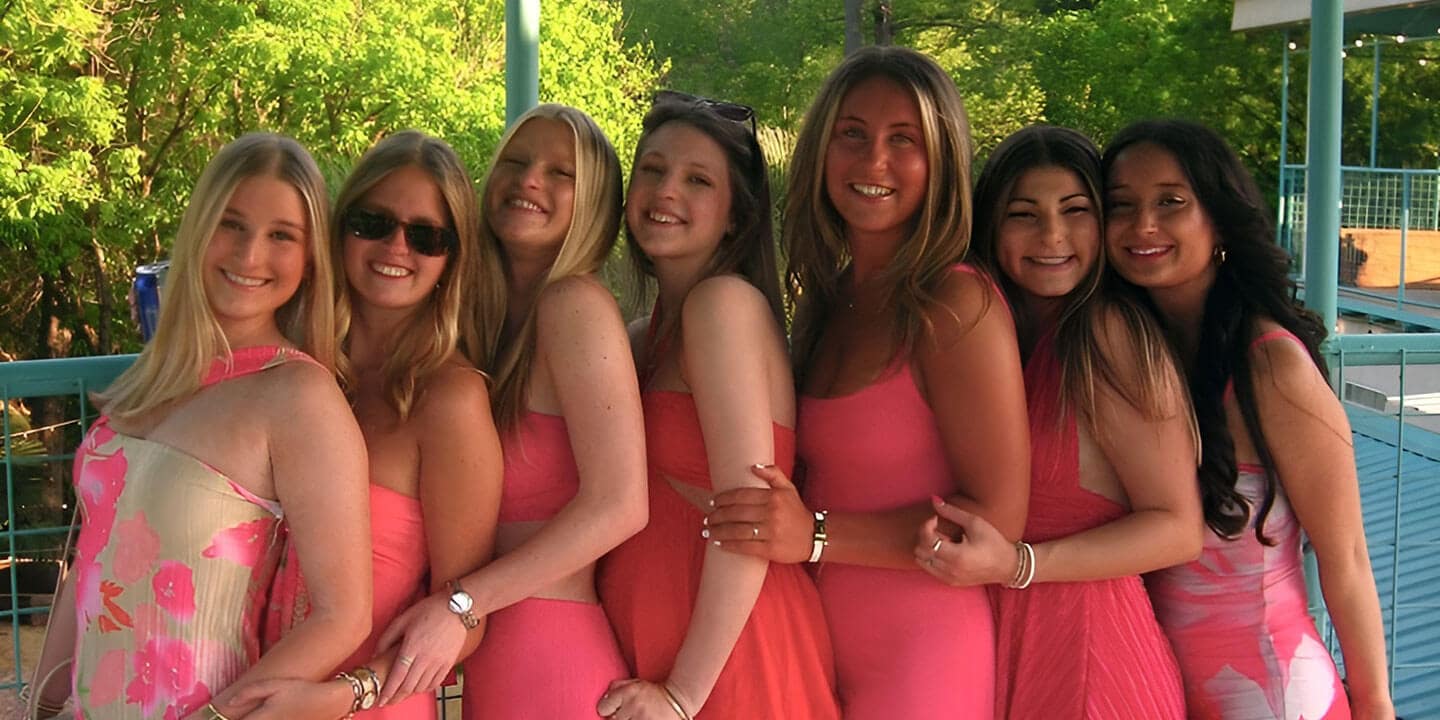 Seven girls standing sideways in a row wearing brightly colored dresses outside.