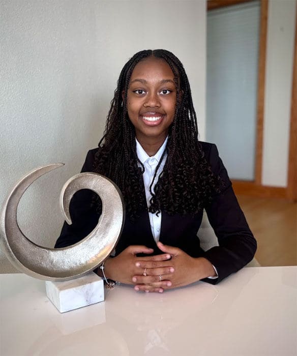 The blog author has long dark braids, is wearing a black blazer with a white blouse, and is seated at a table with a circular art sculpture in front of her.