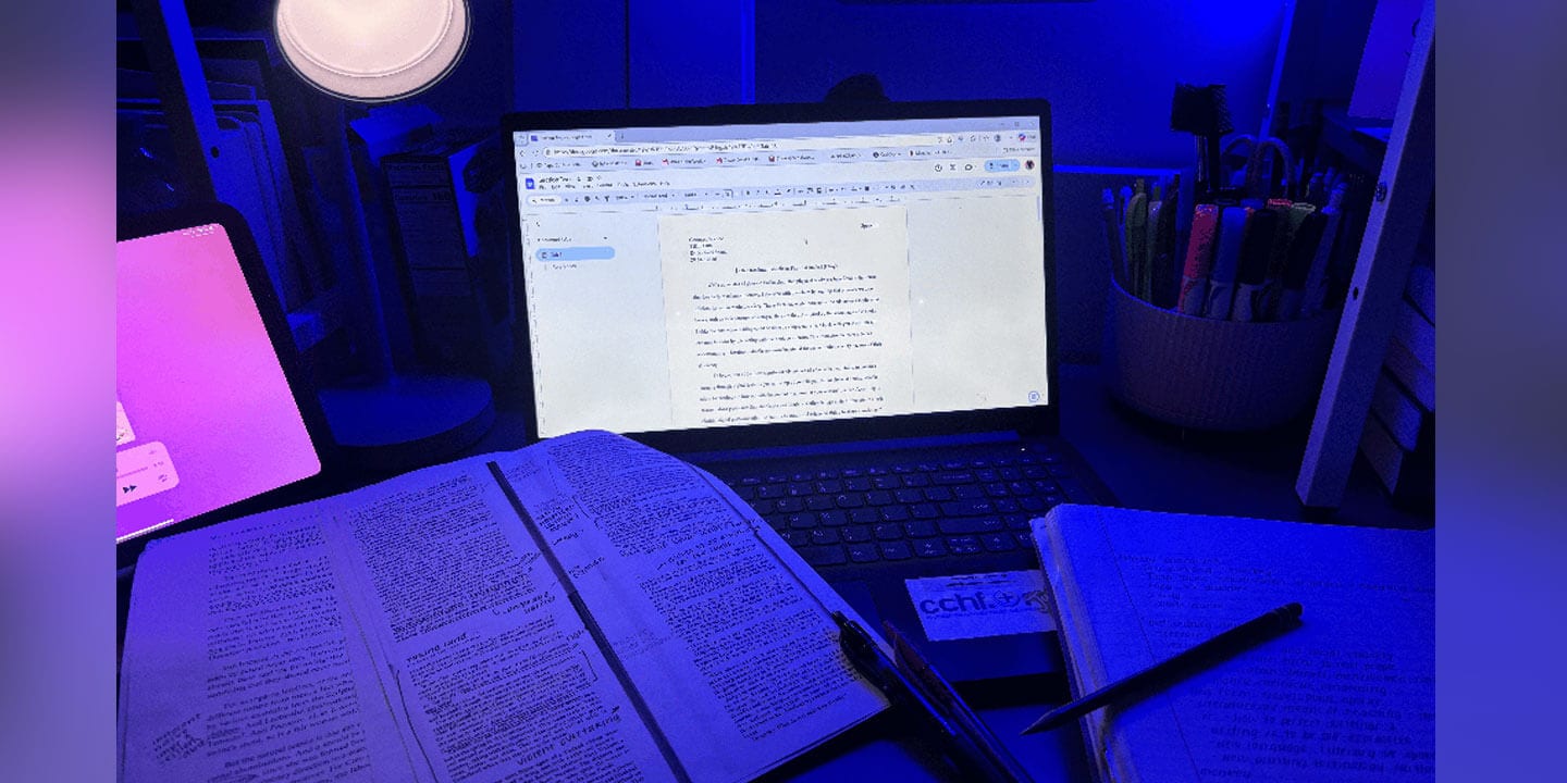 A laptop surrounded by papers, a notebook, a tablet, and pens lit by blue light suggesting a late-night study session.