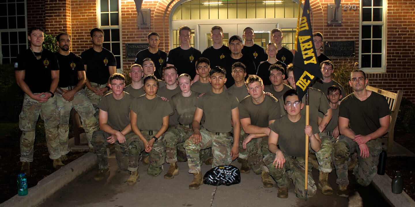A group of Army ROTC cadets in front of a brick building. Some students are wearing black t-shirts and others are wearing army green t-shirts. All cadets are wearing camouflage pants. One cadet is holding an Army flag.