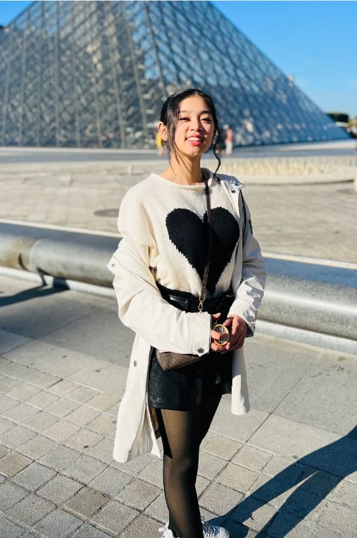 The Blog Author, Denise, is standing in front of the Louvre wearing a white sweater with a black heart on it and a black skirt.