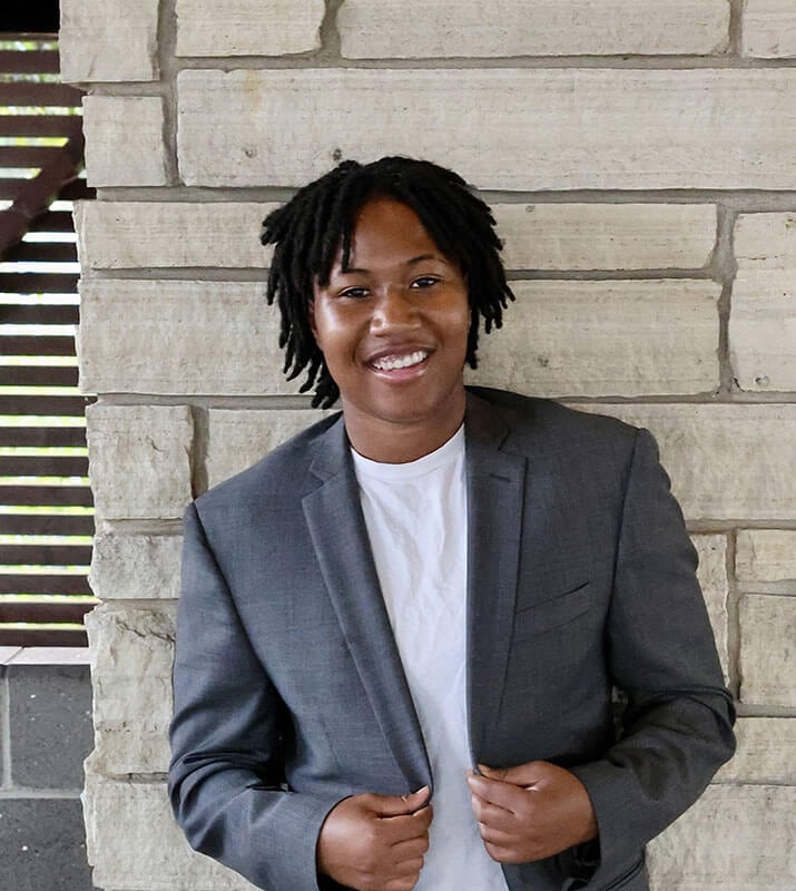 Blog author Dawson has short black braids and is wearing a dark grey blazer over a white t-shirt.