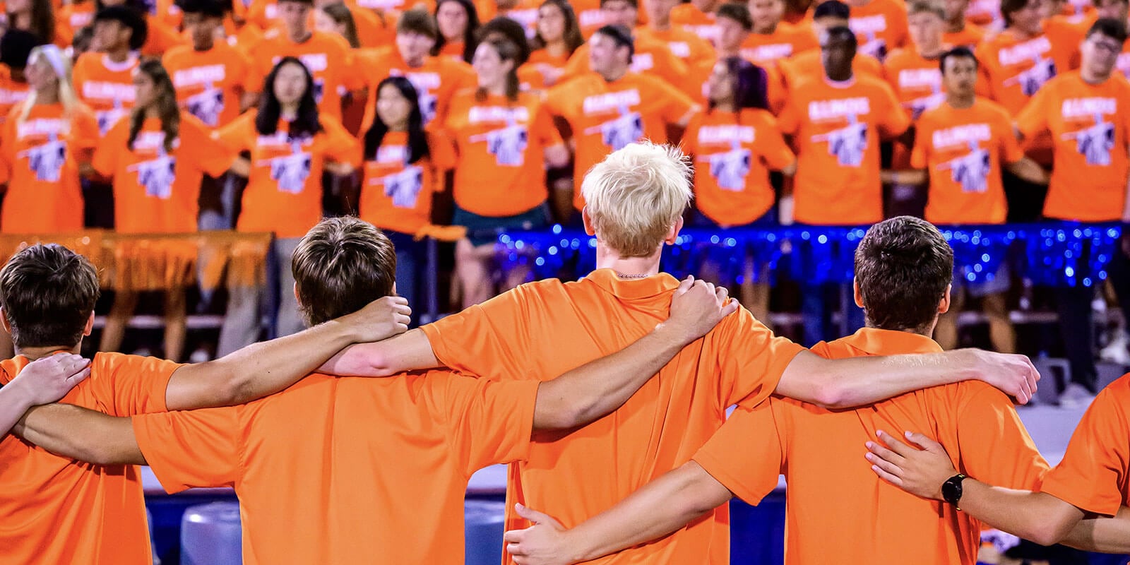 Image shows four teammates with their arms around each other and their backs to the camera facing the crowd cheering them on.