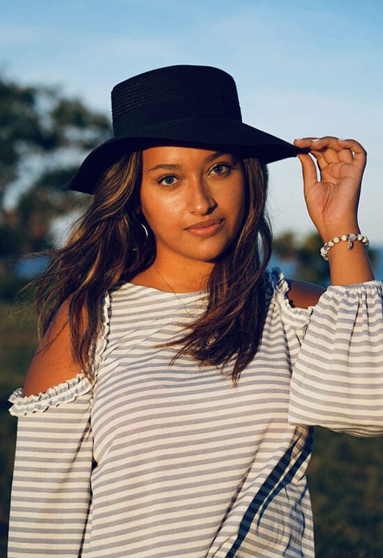 Blog author Jasmine is standing outside on a beach. She is wearing a black fedora-type hat and a striped long sleeve top.