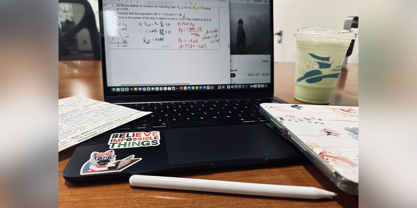 A desk scene showing a laptop with equations pictured, a stylus, a piece of paper with printed notes, and a green drink.