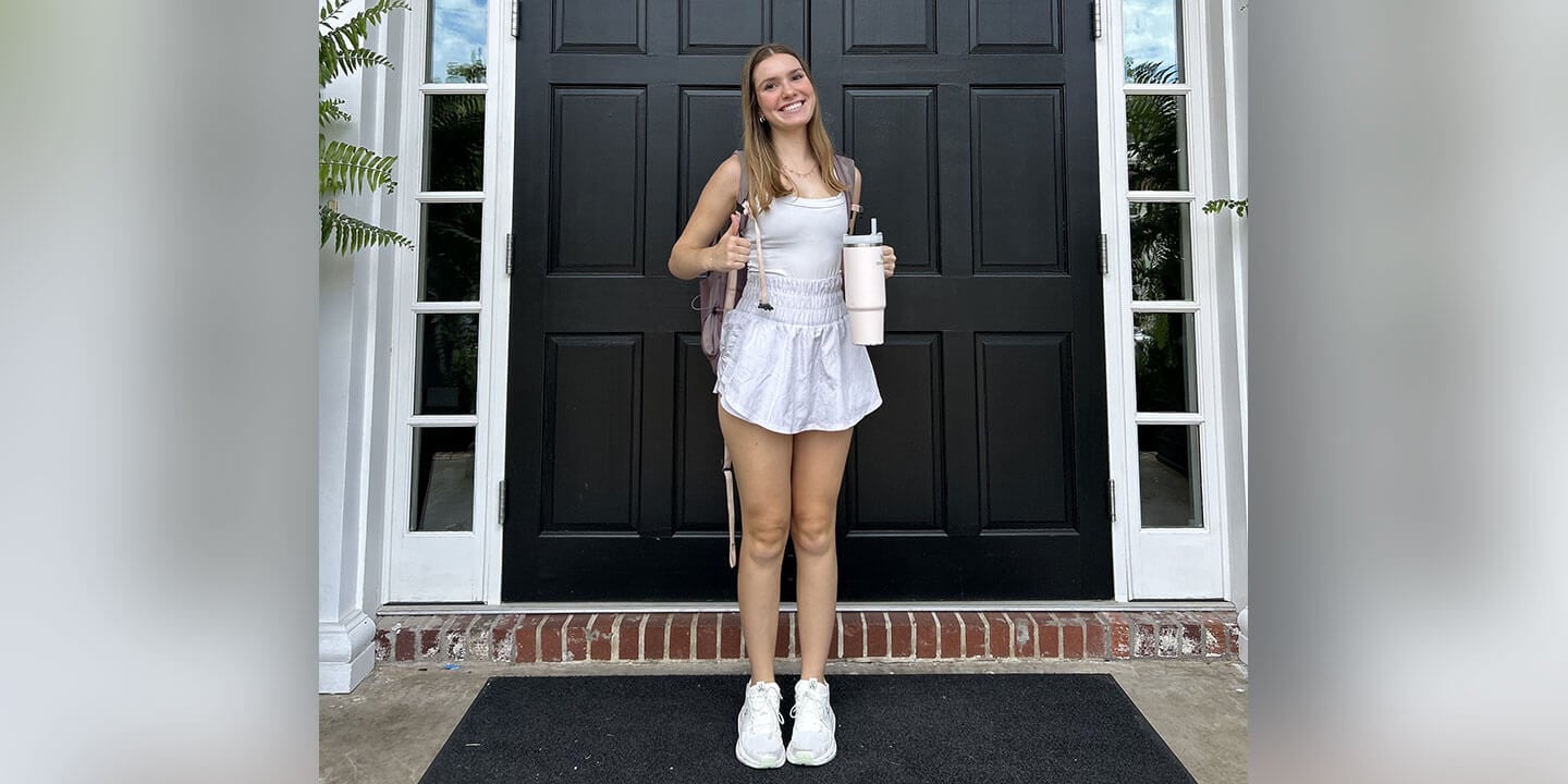 A young college woman standing on her porch with a backpack and large water bottle.