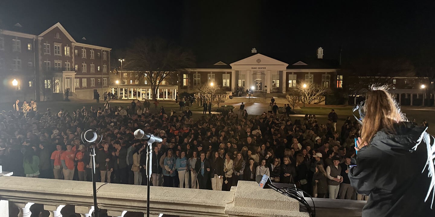 A large group of people gathered outdoors on a college campus at night before a speech or gathering is about to occur.