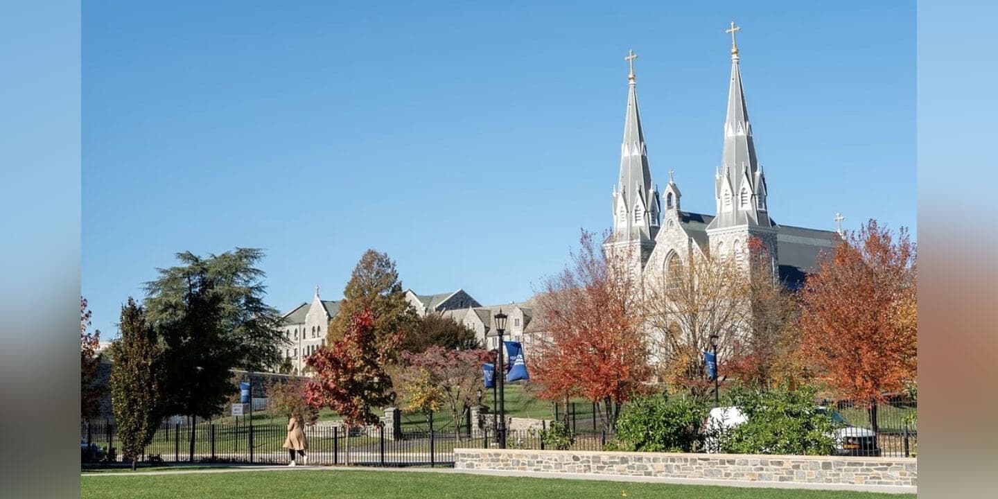 Image shows a large stone Gothic-style church on a college campus in the fall.  A low stone wall, black fence, and a walkway are pictured in front of the church.