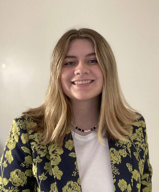 Blog author Jordan has blond hair and is wearing a dark blazer with a yellow floral print over a white shirt.