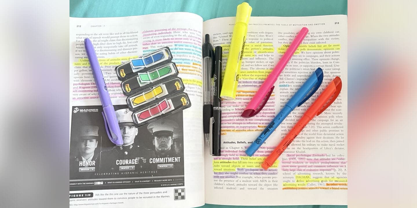An open textbook covered in highlights. There are several highlighters, pens, and colorful sticky tabs lying on top of the textbook.