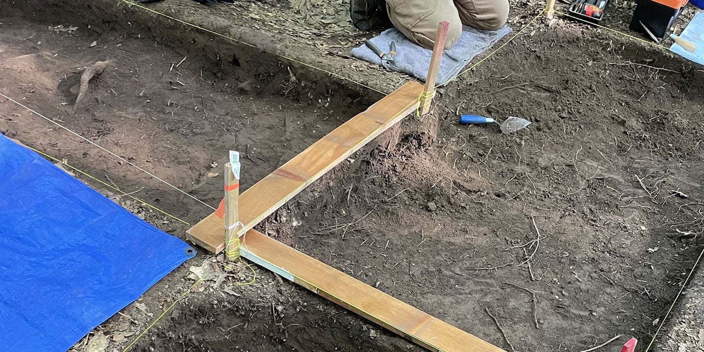 An active archaeological dig site divided into sections by stakes and string with someone kneeling in front of a trowel.