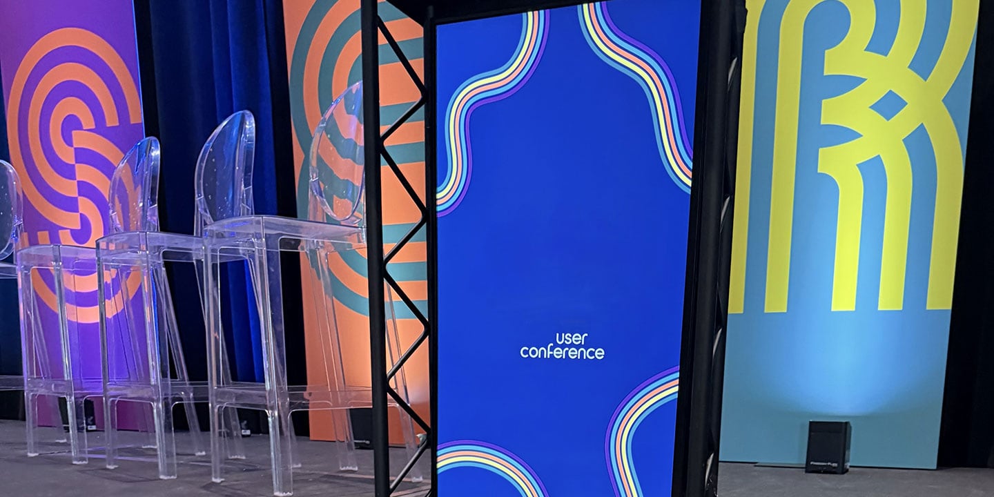 Picture shows a stage with three acrylic stools and a brightly lit sign with the words “user conference” on it.