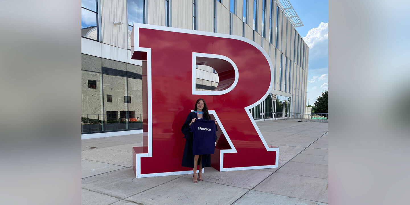 A graduate holding a purple Pearson shirt is standing in front of a large red sculpture of the letter R.