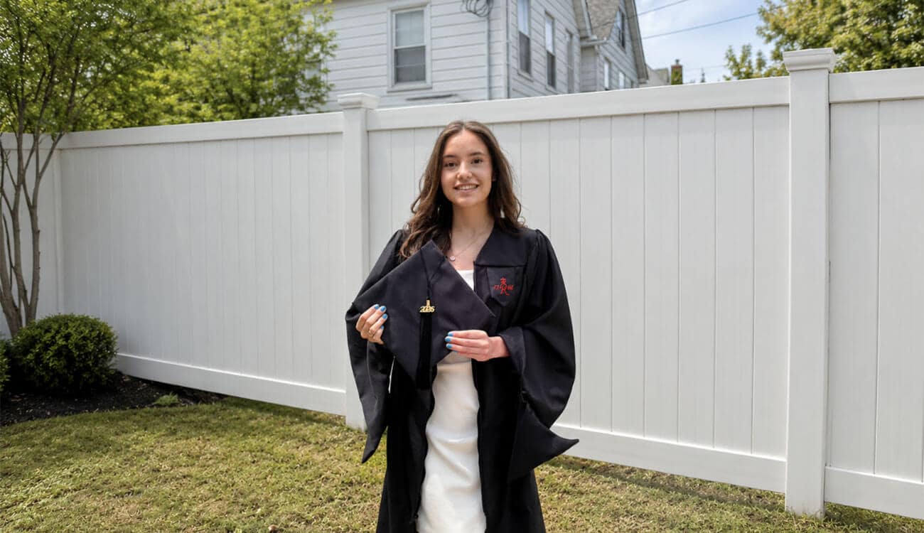A graduate wearing a white dress and an open graduation gown is holding a graduation cap in front of a white fence.