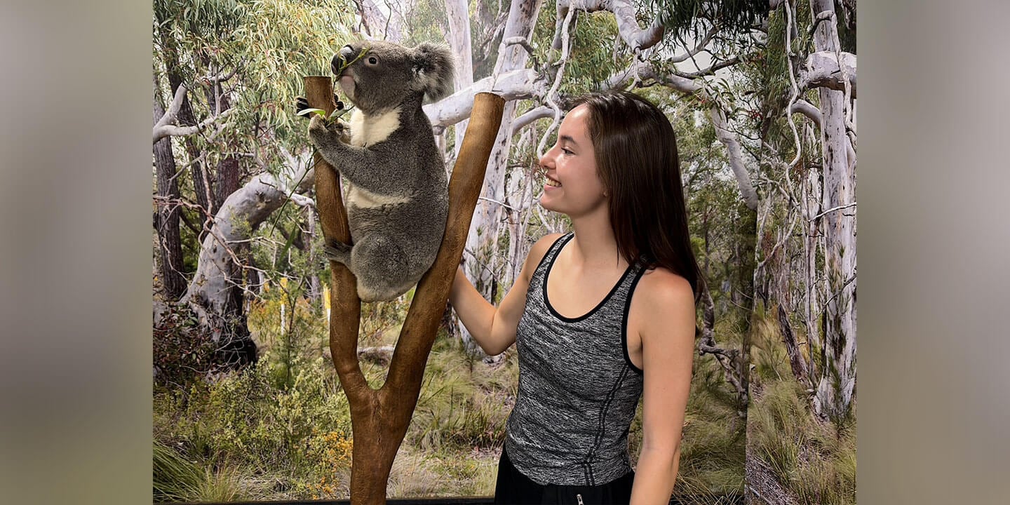 The blog author, Madeline, is wearing a gray athletic tank top and smiling at a koala perched on a tree branch in a wooded area.
