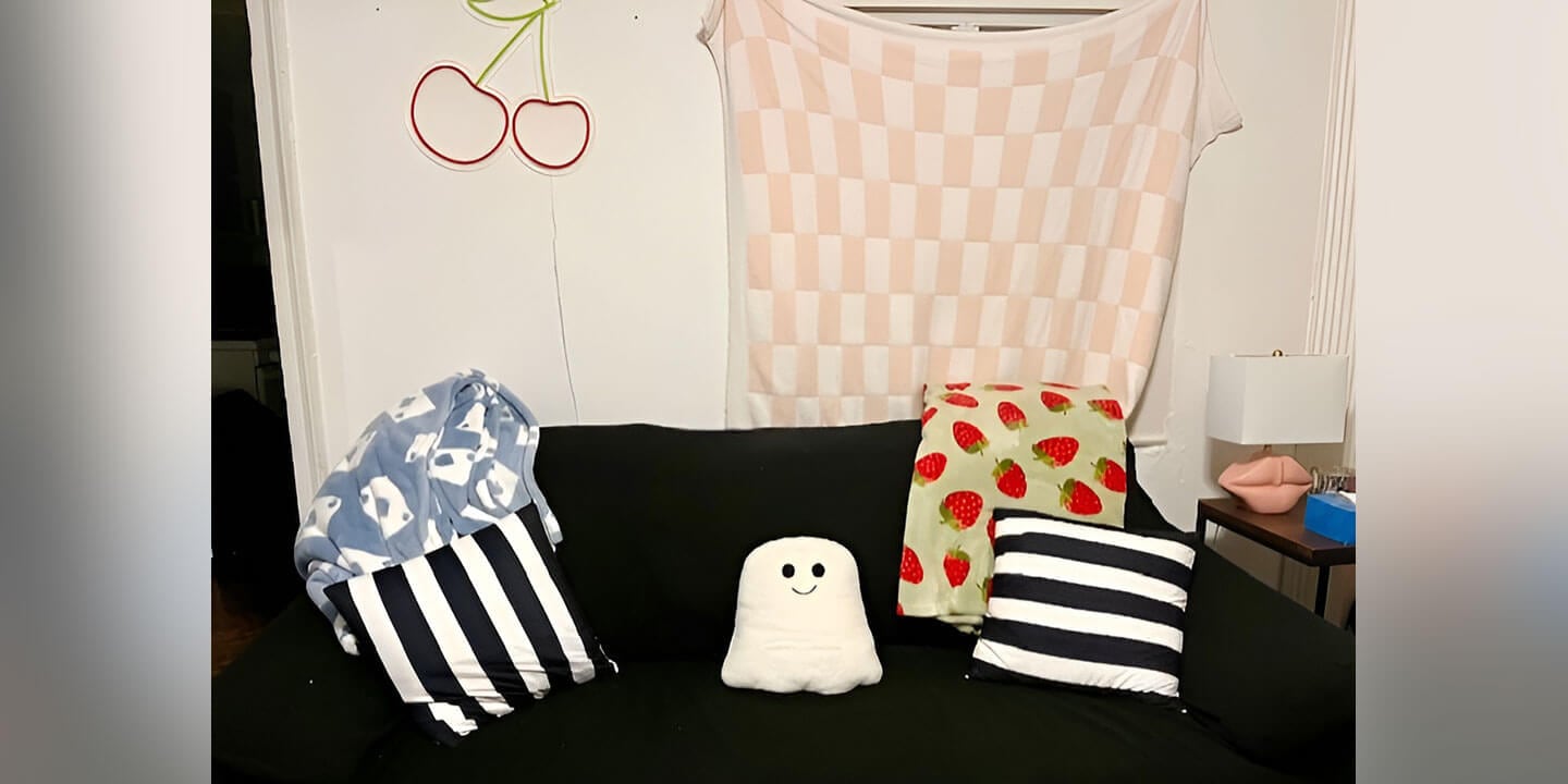 A black couch with two black and white striped pillows, a ghost pillow, a strawberry throw blanket, and a blue and white throw blanket draped over the back.