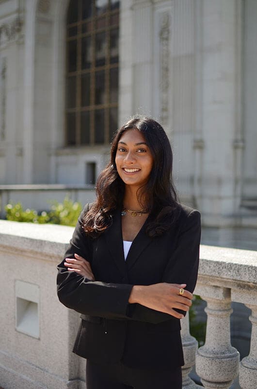 Blog author Mira standing with her arms crossed outside by a concrete railing. She has long dark hair and is wearing a dark suit.