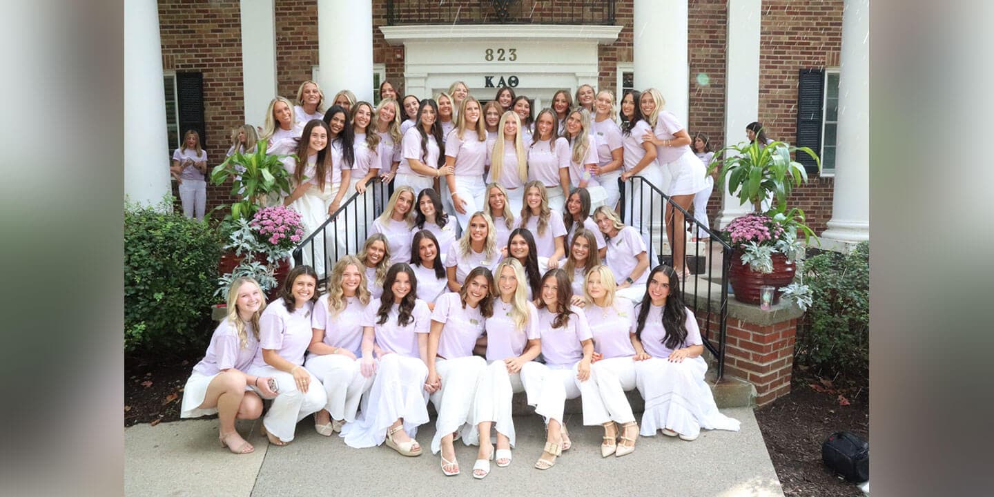 A group of sorority sisters wearing pink shirts and either white skirts or pants gathered together in front of a sorority house.