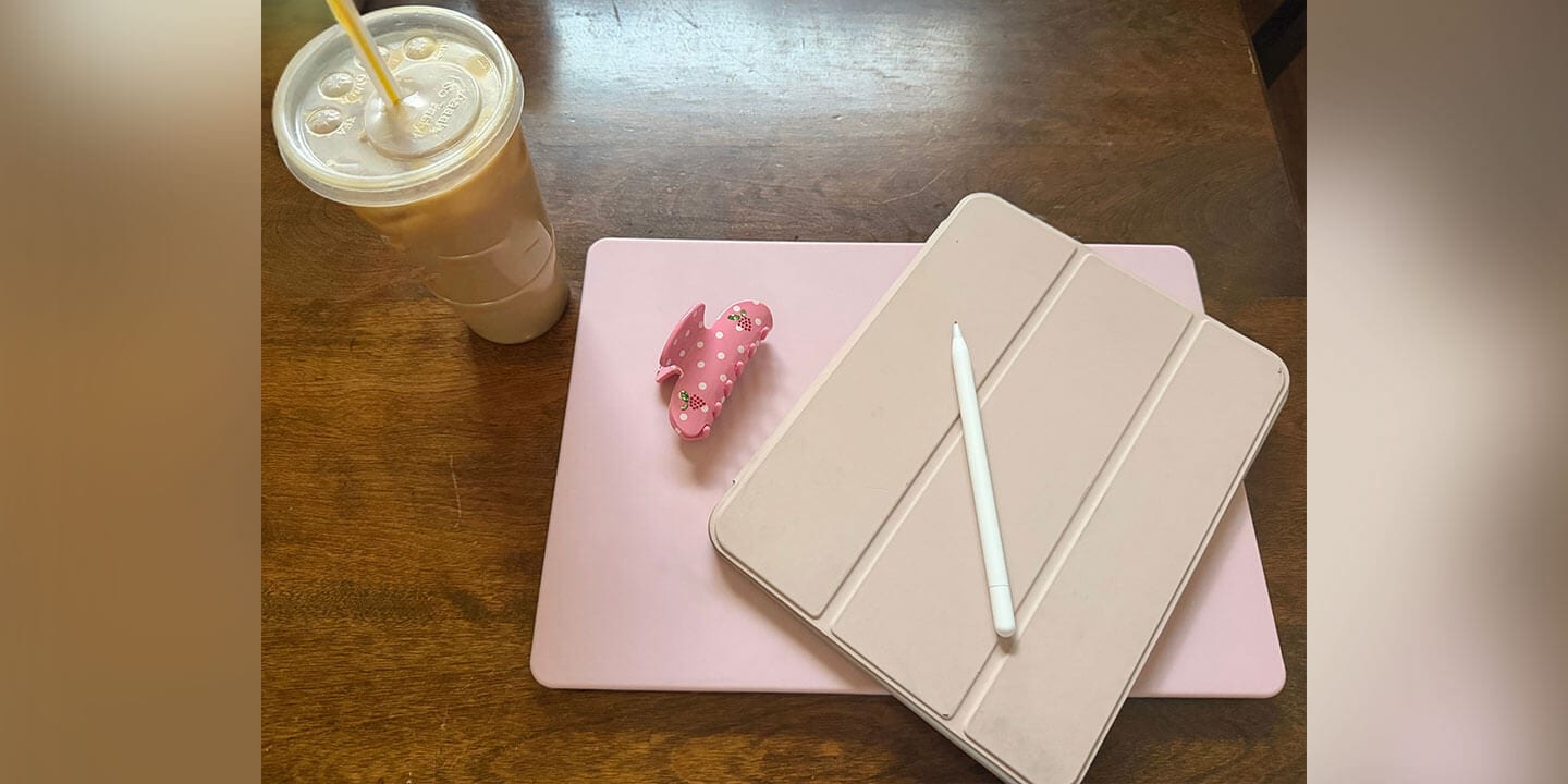 A closed beige iPad with a white stylus rests on a pink pad with an iced coffee and yellow straw placed next to it on a wooden table.
