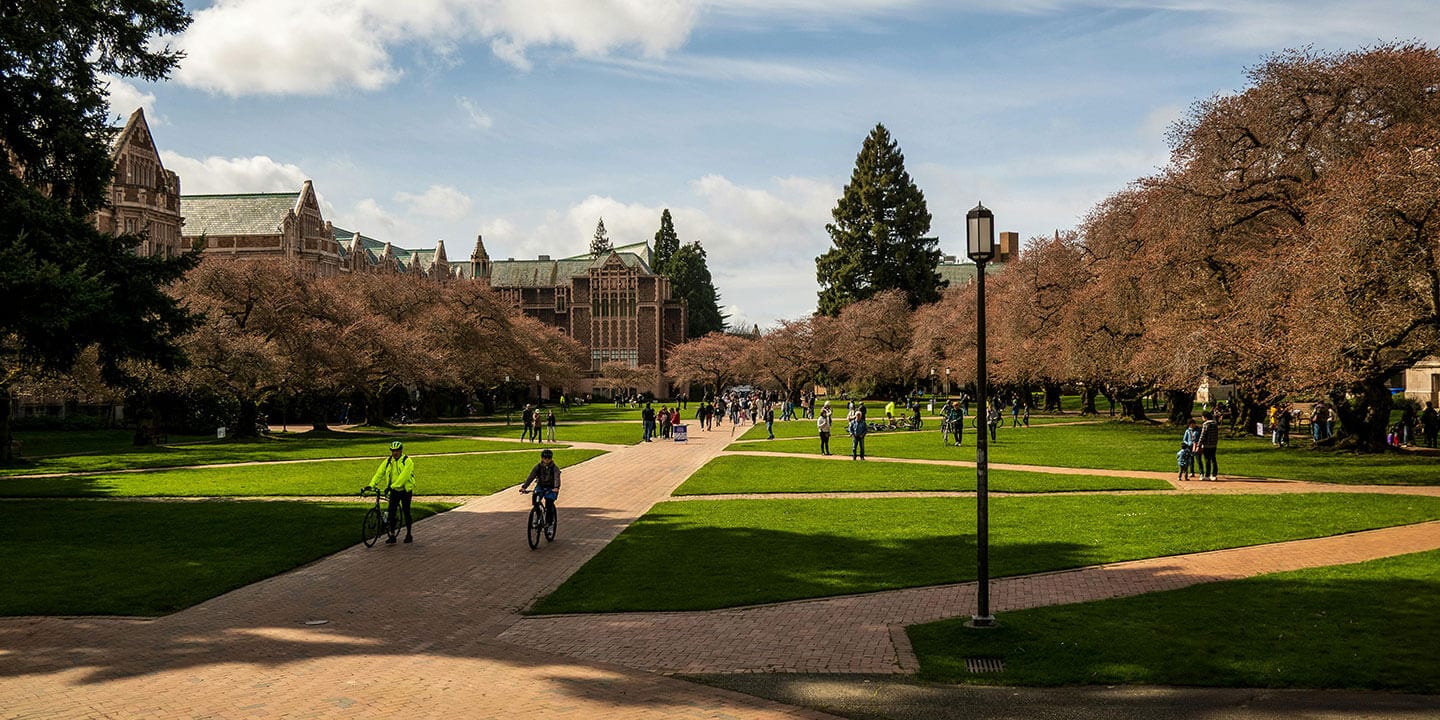 A college campus on a sunny day with students walking and biking across campus in front of historic campus buildings.