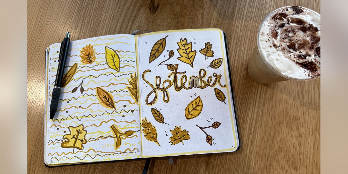 An open journal filled with doodles of fall leaves and the word September and a black pen and an iced coffee drink on a wooden surface.