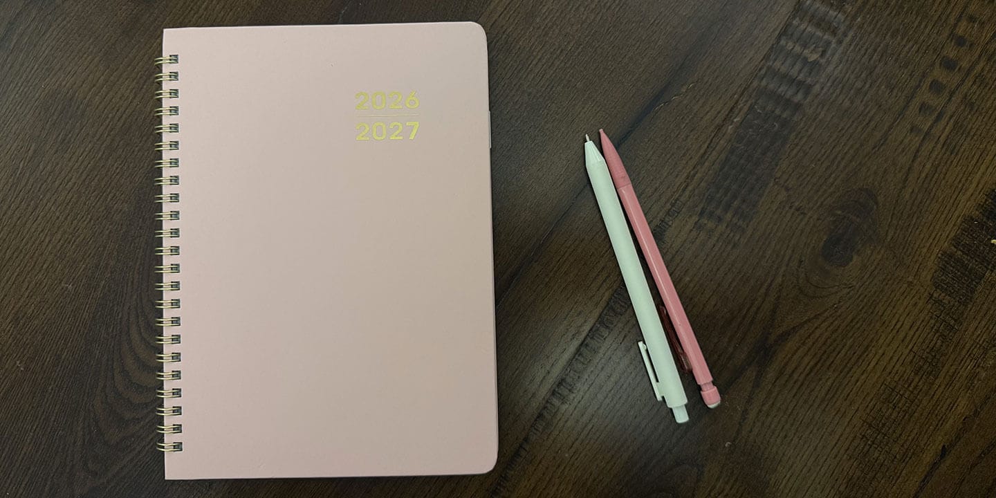 A pink planner pictured on a table with a pink mechanical pencil and a mint green pen lined up next to it.