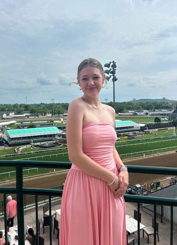 Blog author Riley is wearing a pink strapless dress and standing outside against a railing overlooking a football field.
