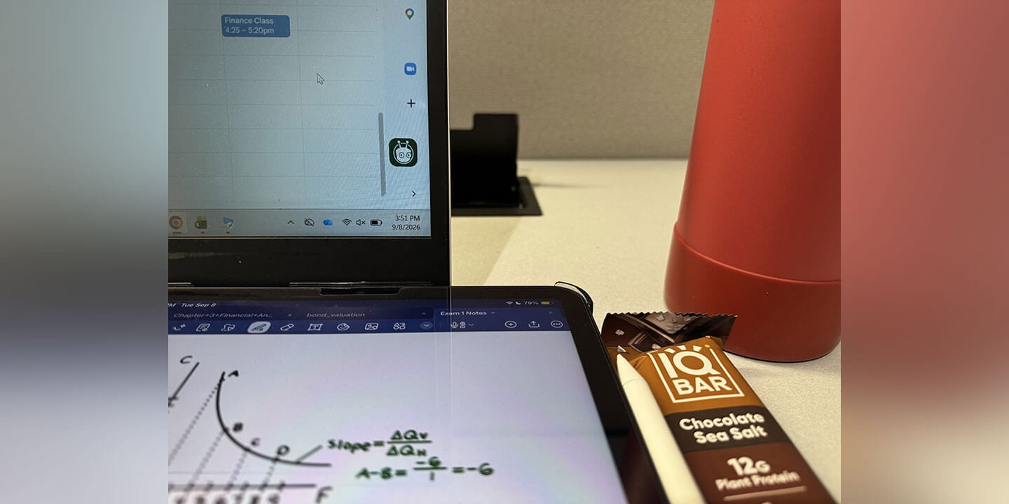 Image shows a desk with a computer screen with a schedule pulled up, an iPad with a graph showing, an energy bar, and a water bottle.