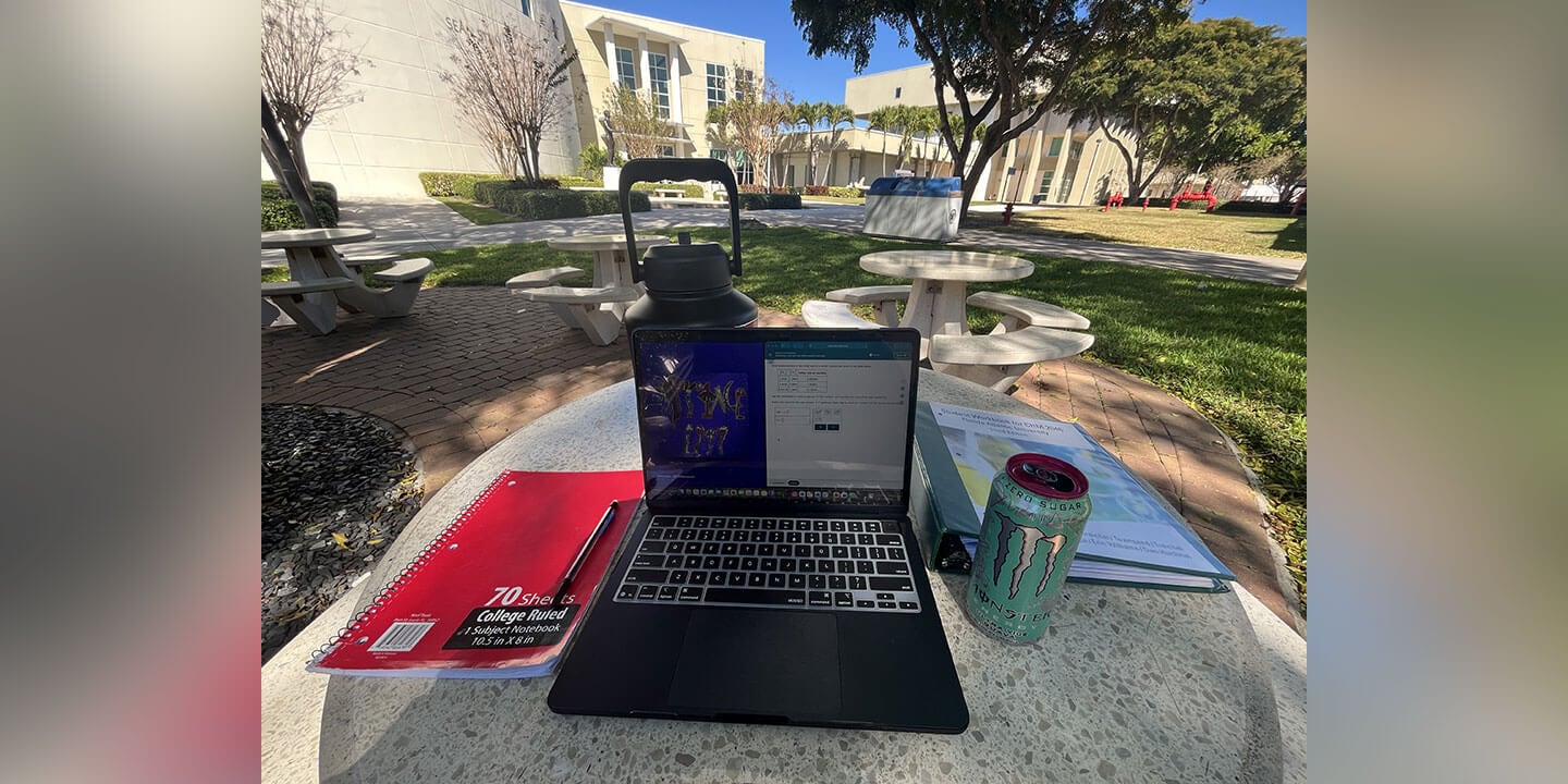 An outdoor study scene showing a laptop set up on a cement table surrounded by a notebook, binder, energy drink, and a water bottle on a college campus