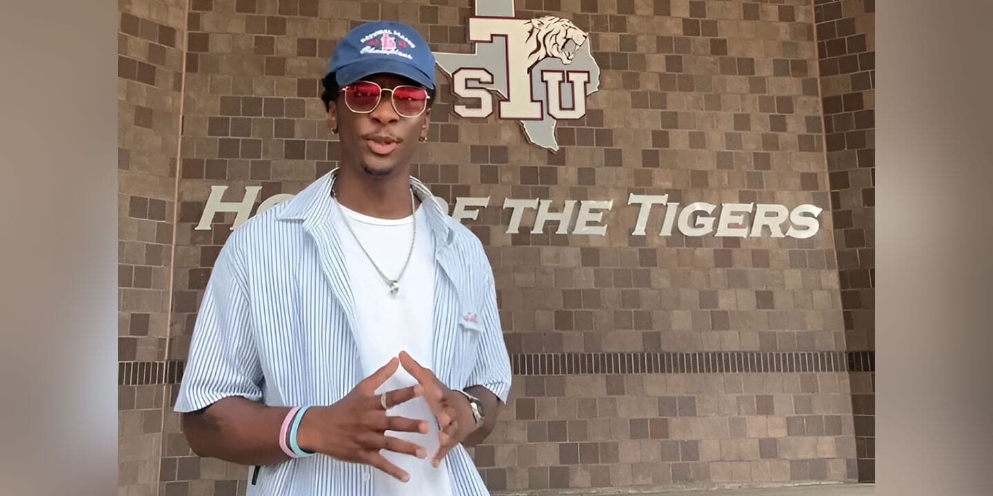 A young man wearing sunglasses, a ball cap, a silver necklace, and a striped button-down shirt with a white t-shirt underneath standing in front of brick wall with a school logo on it.