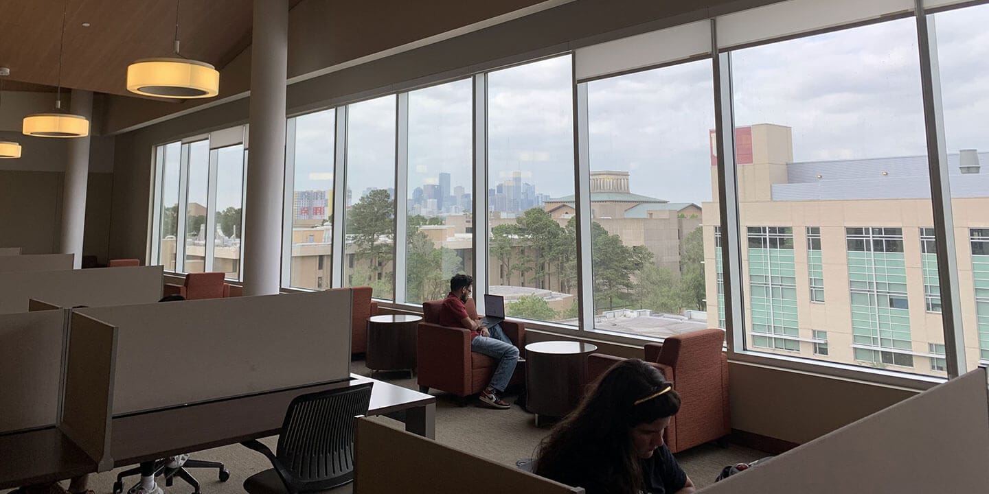 A person wearing jeans and a red polo sitting in an orange chair working on a laptop in front of a bank of windows in a large room.