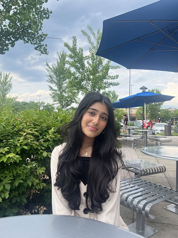 The blog author is seated outside at a patio table under a blue umbrella and has long dark hair and is wearing a cream sweater over a black shirt and a gold half-moon necklace.
