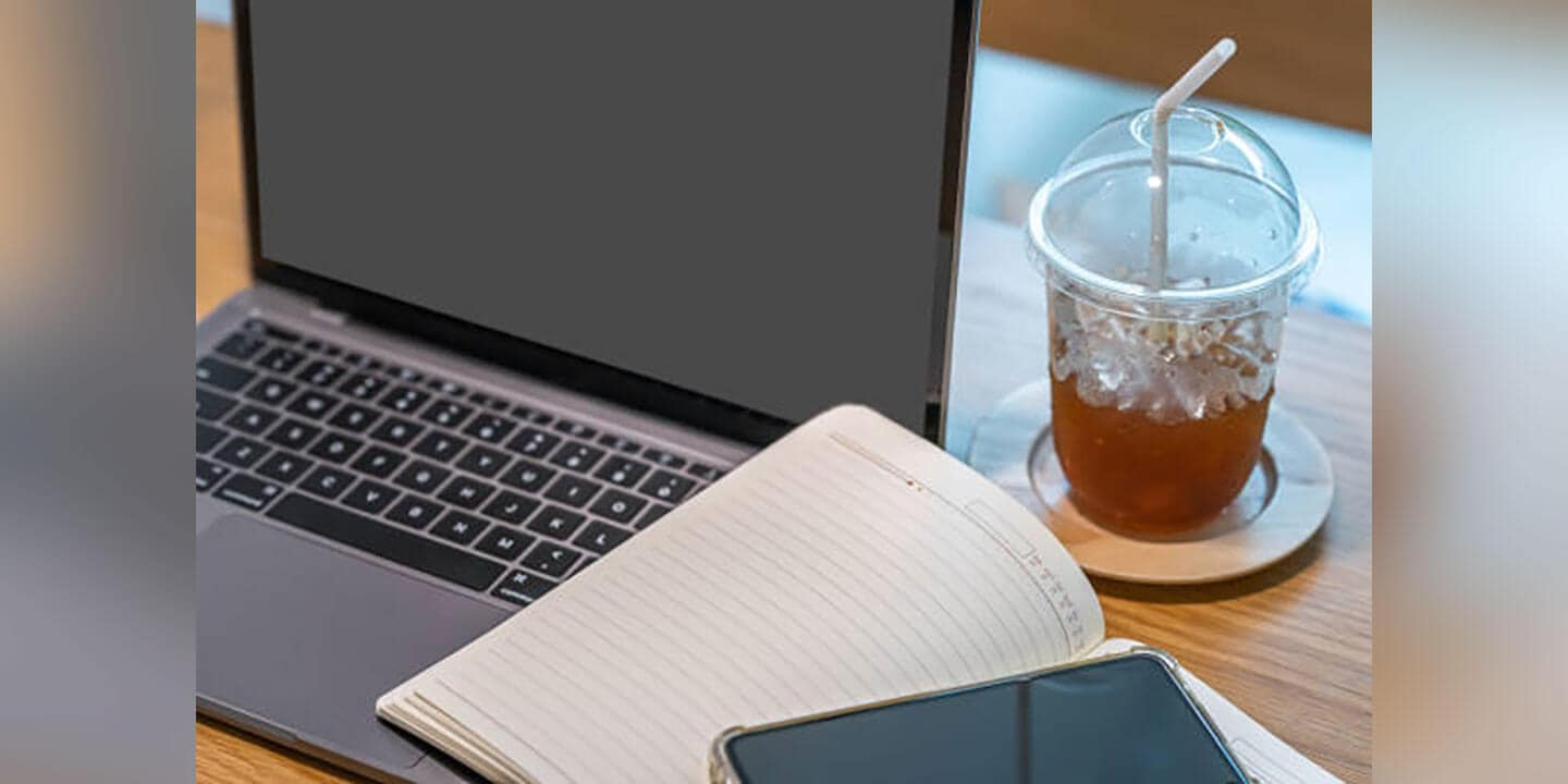 A to-go cup of iced tea, a laptop, a notebook and a phone placed on a desk.