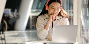 A girl wearing headphones is sitting at a desk, leaning her chin in her hand, gazing at a laptop screen.