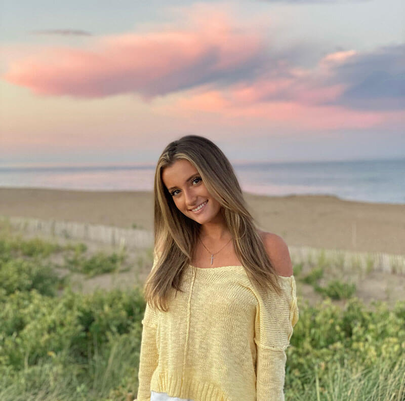 Blog author Ava is standing on a beach with the sunset in the background. She has long light brown hair and is wearing a yellow sweater.