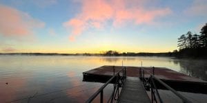 A dock on a lake with the sunset on the horizon. The clouds look pink in the evening light.
