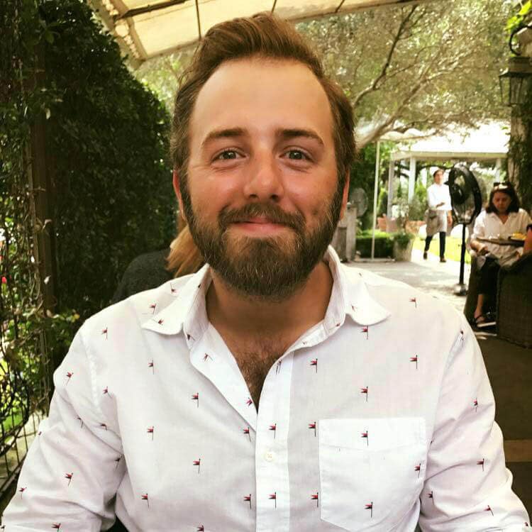 In this profile photo, blog author Andrew Bierbower is smiling and sitting at an outdoor restaurant patio under an awning. He has short brown hair with a full beard and mustache. He is wearing a white collared shirt with a small black pattern.