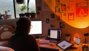 A young female college student sits at a desk in her room working on a laptop computer. There is also a desktop monitor and tablet open on her desk. There are various posters on the wall in front of her, including one for Harry Styles.
