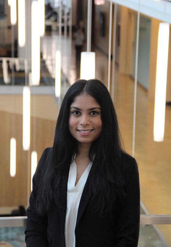 In this profile picture, blog author Ankita Chittiprolu is smiling and standing in front of some hanging light fixtures. She is a young female college student with long dark hair and is wearing a dark blazer over a white blouse.
