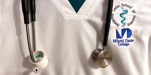A v-neck scrub shirt with a stethoscope around the neck. The pocket logo says, “Benjamin Leon School of Nursing, Miami Dade College”.