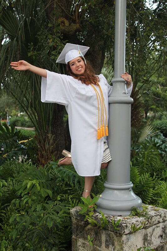 Blog author Ana Cooper is wearing a white cap and gown with two gold honor cords. She is perched on a lamp post and holding out her right arm.
