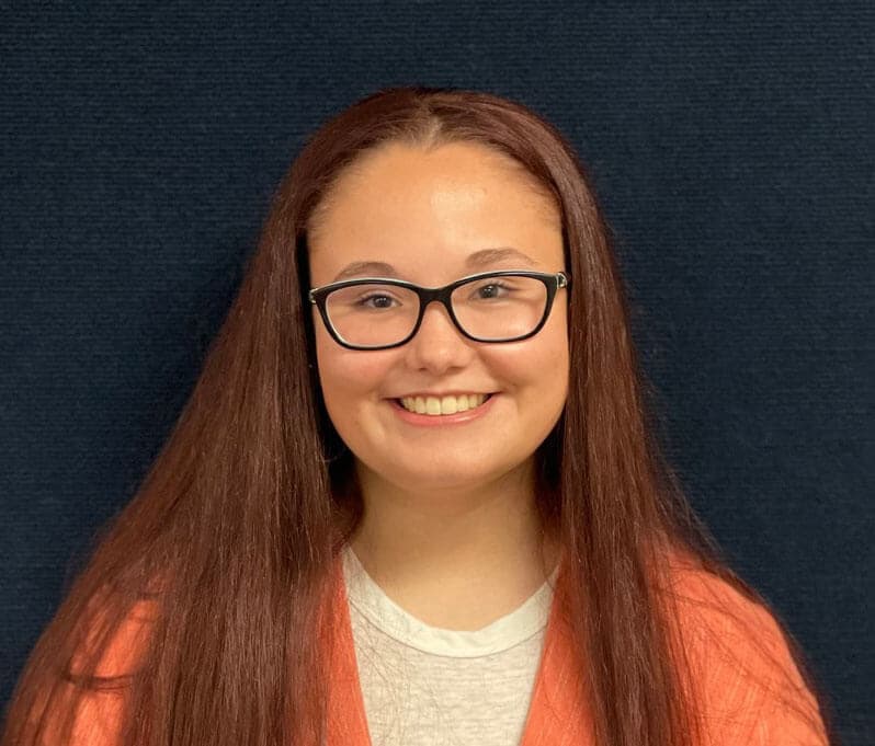 Blog author Abigail has long dark brown hair and is wearing glasses. She is standing against a dark background and is wearing an orange cardigan over a white shirt. 