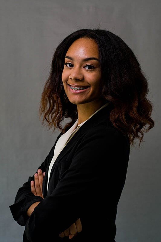 Blog author Ashanti is smiling and standing in front of a grey neutral backbround with her arms crossed. She is wearing a dark blazer over a white blouse.