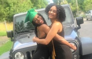 Two female high school students are smiling and hugging in front of a Jeep vehicle. They are both wearing black dresses and the female on the left is wearing a green graduation cap.