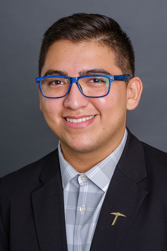 Blog author Angel in a studio headshot. He has short dark hair and is wearing blue horn rim glasses, a dark suit jacket over a light grey checkered shirt. 