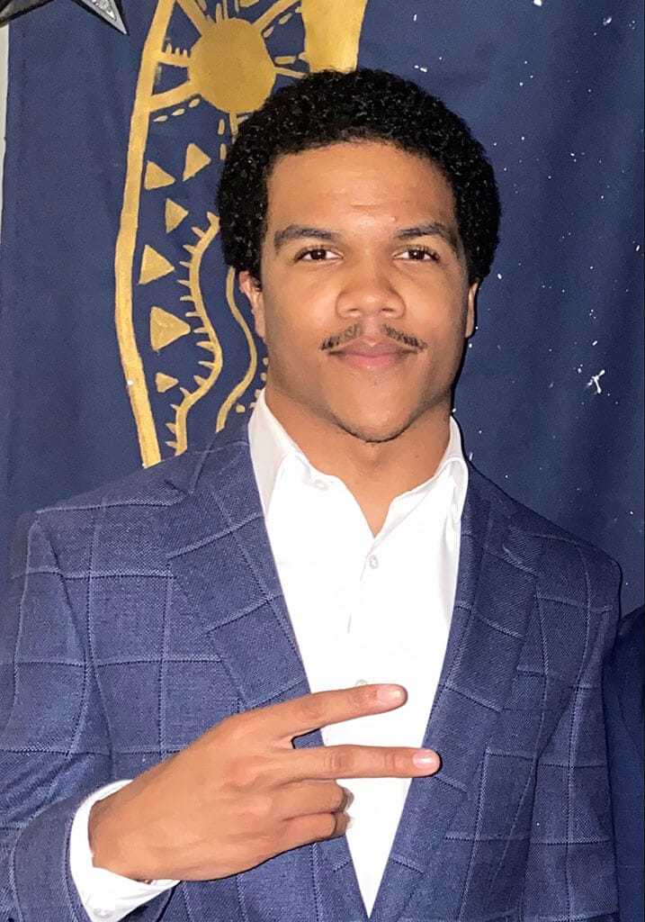 Blog author Brian Hayes is standing in front of a navy blue background with gold lettering. He has short dark hair, is wearing a blue sport coat and white shirt, and is holding up two fingers on his right hand.