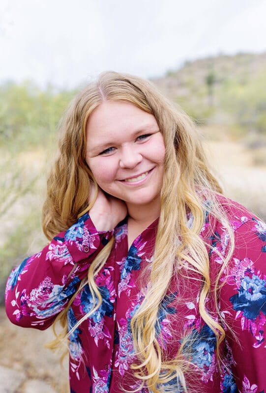Blog author Bethany Robinson smiles and stands outside on a nature trail. She has long blonde hair and is wearing a purple blouse with blue flowers.