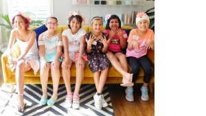 Six young female cancer patients are smiling and sitting on a couch. They are wearing make-up and dress-up clothes.